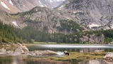 Tranquil mountain lake surrounded by rocky peaks with patches of snow and dense forest, with a black dog standing near the water on grassy land