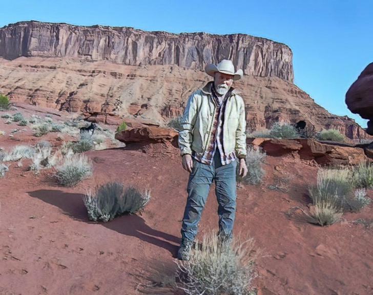 Man wearing a cowboy hat and layered clothing standing on red rocky desert terrain with sparse desert vegetation and a large flat-topped mesa in the background under a clear blue sky