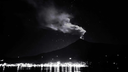 Volcano at night emitting smoke and sparks above a lit shoreline with reflections on the water