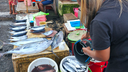 Two people at a fish market with fresh fish displayed on a table and in buckets, with one person pouring fish from a bucket