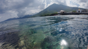 Calm clear water revealing coral formations beneath the surface, with a mountainous coastline and scattered buildings under a partly cloudy sky in the background