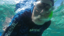 Person snorkeling underwater wearing a dark wetsuit, diving mask, and snorkel, reaching out towards the camera in clear blue water