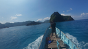 Person standing at the front of a wooden boat on vibrant blue water headed toward lush, green islands under a clear sunny sky