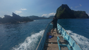 Person standing at the bow of a wooden boat with blue sides, heading towards lush green islands under a bright blue sky over calm ocean waters
