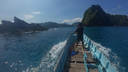 Person standing at the front of a small blue boat navigating toward rocky islands under a clear blue sky over calm sea water