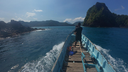 Person standing at the bow of a wooden boat moving through blue water near rocky and forested islands under a clear sky