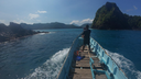 Man standing at the front of a small wooden boat heading toward a lush, rocky island under a clear blue sky