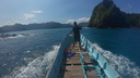 Person standing on the wooden bow of a small boat taking a photo of a large steep green-covered island surrounded by blue ocean waters under a clear sky