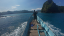 Person standing at the bow of a wooden boat navigating through bright blue water towards a forested rocky island under a clear sky