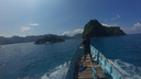 View from a boat moving through clear blue ocean waters toward green, rocky islands under a bright blue sky with scattered clouds, with a person standing at the boat's bow looking ahead