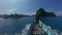 Person standing at the front of a small wooden boat heading toward lush green rocky islands under a clear blue sky