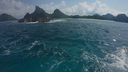 Turquoise sea with gentle waves in the foreground and a rocky, forested island with sandy shores under a bright blue sky with scattered clouds in the background