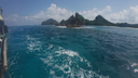 View of clear blue ocean water with frothy waves near a boat, rocky islets, and forested islands under a partly cloudy sky