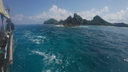 View of vibrant turquoise ocean water with gentle waves, rocky islets covered in greenery, and a partly cloudy blue sky, seen from the side of a wooden boat