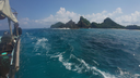 View from a boat on clear blue ocean waters approaching a small island with rocky, forested hills under a partly cloudy sky