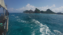 View from a boat on clear blue water approaching a lush green island with rocky, jagged hills under a partly cloudy sky