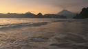 Waves gently rolling onto a beach at dusk with silhouetted volcanic mountains in the distance under a softly glowing sky