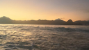 Sunset over a calm sea with waves gently rolling towards the shore and a silhouette of distant mountain peaks on the horizon