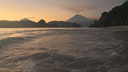 Gentle ocean waves rolling onto a sandy beach at sunset with a distant smoking volcano silhouetted against a warm sky