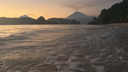 Calm ocean water gently washing onto a sandy beach with a silhouette of a smoking volcano and nearby hills at sunset