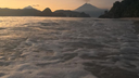 Foamy waves washing onto a beach at sunset with a volcanic mountain and hilly landscape in the background under a softly colored sky