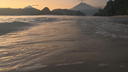 Calm ocean waves gently rolling onto a sandy shore with a silhouette of volcanic mountains under a soft evening sky