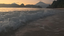 Waves gently crashing onto a sandy shore with silhouetted volcanic mountains under a colorful sunset sky