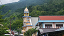 A quiet village street lined with houses and a church tower with a golden dome against a backdrop of lush green forested hills under a cloudy sky