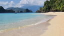 gentle waves lapping on a sandy beach with lush green cliffs on the right and rocky islands in the distance under a partly cloudy sky