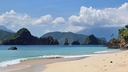 Sandy beach with gentle waves lapping the shore, clear turquoise water, and green, rocky islands under a partly cloudy blue sky
