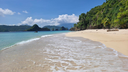 Clear water gently washing onto a sandy beach with green, forested hills rising on the right and distant rocky islands under a blue sky with scattered clouds
