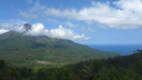 Volcanic mountain partially covered by clouds above lush green forest with a distant view of the ocean under a partly cloudy blue sky
