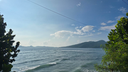 Calm sea waves rolling gently toward a shoreline framed by green foliage under a bright blue sky with scattered clouds and distant mountainous islands on the horizon
