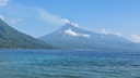 Calm blue sea in the foreground with a large, green forested mountain rising behind it, topped by a volcanic peak emitting a small plume of white smoke under a clear blue sky