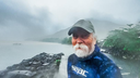 man with white beard and cap wearing a dark blue Seac wetsuit standing on a rocky shoreline with misty mountains and water in the background