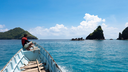 Man sitting at the bow of a wooden boat navigating through blue water toward green, rocky islands under a bright sky with scattered clouds