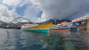 Colorful boats floating on clear water near a dock with buildings and a mountain partially covered by dark clouds in the background