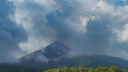 Distant volcano peak partially obscured by large clouds with forested hills in the foreground