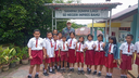 Group of school children in red skirts and shorts with white shirts standing outside a school building under a sign with Indonesian text