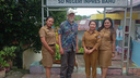 Four people standing together outdoors in front of a sign with Indonesian text, three women wearing matching tan uniforms and one man wearing a colorful shirt and gray pants
