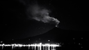 Volcano erupting at night with glowing lava, smoke plume rising, starry sky, and lights from a nearby settlement reflected on calm water