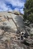Person sitting on rocky terrain beside a black and white dog, with large boulders and pine trees in the background under a partly cloudy sky
