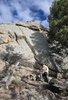 Man wearing outdoor gear and a hat sits on a rock at the base of a large, sunlit granite cliff surrounded by trees and dry grass under a partly cloudy blue sky