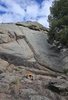 Large rock formation with rough textured surface under a partly cloudy sky, a small fire burning on the ground among rocks and dry grass, and an evergreen tree partially visible on the right