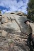 Man wearing a cowboy hat and beige jacket standing next to large rocky boulders under a partly cloudy blue sky with trees around