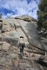Person wearing a wide-brimmed hat and light jacket walking uphill toward a large rock formation under a partly cloudy blue sky, with a small fire burning at the base of the rocks and a dog partially visible on the right
