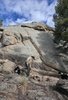 Person in outdoor clothing tending a small fire near large rocky boulders with two black and white dogs nearby under a partly cloudy sky
