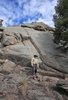 Man wearing a beige jacket and black cowboy hat stands on rocky terrain in front of a large rock formation under a partly cloudy blue sky