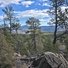 Rocky forest landscape with tall pine trees under a blue sky with scattered white clouds, overlooking distant forested hills and mountains