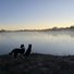 Two border collies sitting by a calm riverbank at dawn with mist rising over the water and a clear sky above